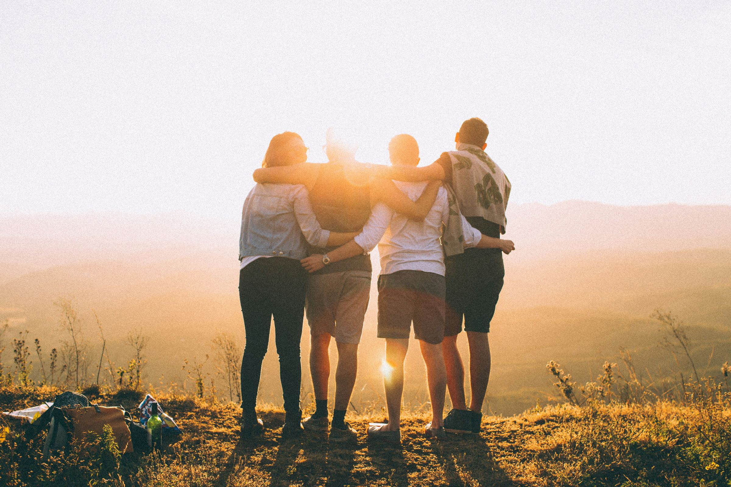 Four friends with their arms around each other's shoulders, watching the sunset over the hills